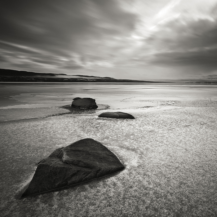 Arrival of Winter, photography by Anders Naesset. The winter arriving. Ice begins to encapsulate the boulders by the lake shore. Snowshowers lurks overhead.. Canon EOS 5D, Canon EF 17-40L, Hoya ND400, Cokin Z-Pro ND8 graduated grey. Toned. In Nature, Scenery, Waterscape
