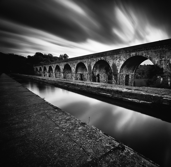 The 70 feet high Chirk aqueduct was built between 1796 and 1801 by Thomas Telford and William Jessop to carry the Ellesmere Canal. The aqueduct is situated on the border of Wales and England.
The ten circular masonry arches of the structure, each spanning 40 feet, have piers carried between them as pilaster strips. There is no cast iron trough carrying the water of the canal, as at Pontcysyllte. Instead, the bed is of iron plates, bolted together, with the side walls built of stone quarried locally at Pont Faen (http://www.chirk.com/aqueduct.html).
The viaduct, higher than the aqueduct, is an active railway viaduct and has a double track. It is absolutely exceptional to watch the canal boat and the train pass across these magnificent stone structures.
. ND 3.0. In Construction, Edifice, Bridge. Chirk Aqueduct, photography by Zeiko. Image #398738
